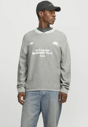 Un pull gris à manches longues avec une texture côtelée, ornée de texte et de patchs blancs. Associé à un jean bleu clair et une casquette noire.
