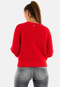 Donna con lunghi capelli castani raccolti in una coda di cavallo, indossa un luminoso maglione rosso a maglia e jeans grigi sbiaditi, vista da dietro.
