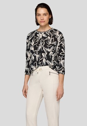 Schwarzes Langarmoberteil mit weißem Blumenmuster, Rundhalsausschnitt und leichtem Stoff. Kombiniert mit cremefarbenen Jogginghosen mit Kordelzug.