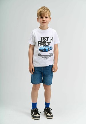 White T-shirt with black text and blue car graphic, paired with denim shorts. Black sneakers and blue socks complete the outfit.