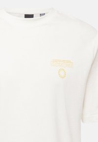 Hvid bomulds-t-shirt med et lille gult grafisk design, der inkluderer ordet "UNKNOWN" og et solstråle-design på brystet.