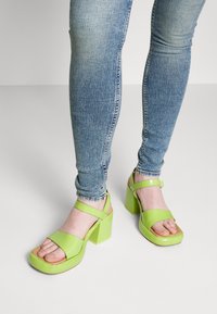 Zelené lakované platformové sandály s blokovými podpatky, s dvěma širokými pásky přes nárt a páskem na patě pro podporu.