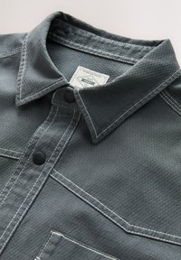 Camicia grigia con texture a motivo di diamanti, dotata di colletto classico, bottoni neri e cuciture a contrasto bianche. Etichetta visibile all'interno del colletto.