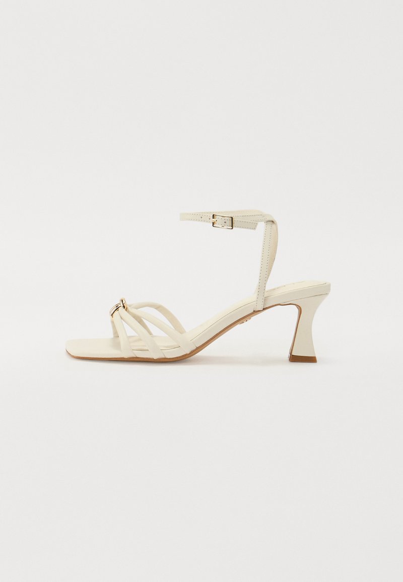 Sandale blanche à talons hauts avec des fines brides, un embout carré et une bride de cheville texturée. Elle présente un talon géométrique et des garnitures en métal doré.