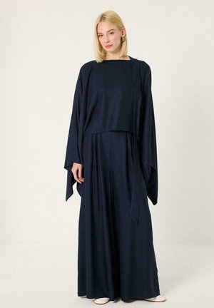 Femme aux cheveux blonds et raides portant une robe bleu marine ample à manches longues et des chaussures blanches, debout sur un fond clair uni.