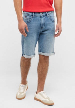 Mann, der hellblaue, hochgekrempelte Jeansshorts, beige-weiße Turnschuhe und ein rotes Hemd trägt, steht vor einem schlichten weißen Hintergrund.