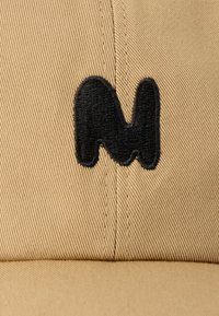 Gorra de tela color caqui con una forma abstracta bordada en negro centrada encima de la visera.