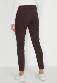 Hnědé slim-fit kalhoty s hladkým povrchem a dvěma zadními kapsami, doplněné světle zbarvenými sportovními botami.