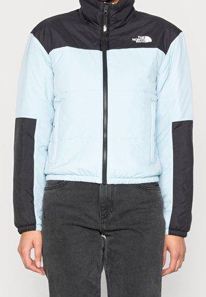 Person iført en lyseblå og sort North Face dunjakke, som er lynet op, med mørkegrå bukser, stående mod en ensfarvet baggrund.