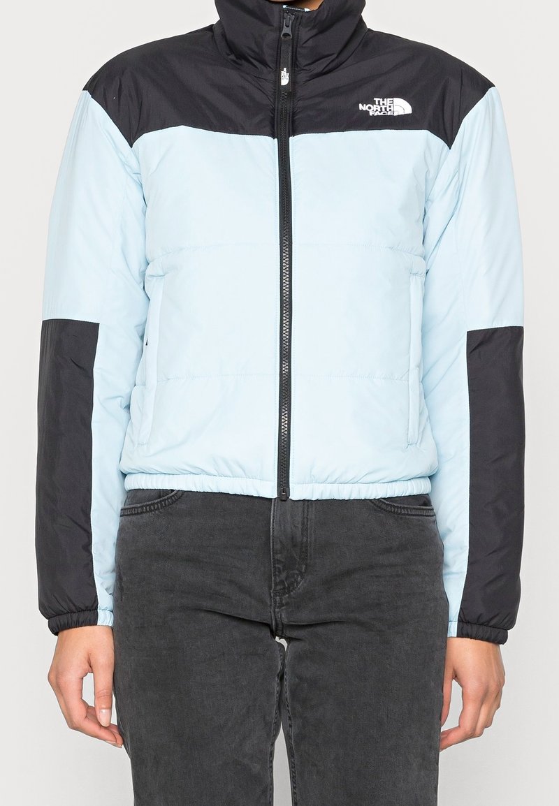 Personne portant une doudoune North Face bleu clair et noir, zippée, avec un pantalon gris foncé, debout devant un fond uni.