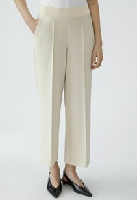Beige, wide-leg trousers with a textured waistband featuring the word "OU" embossed. Designed with front pleats and side pockets.