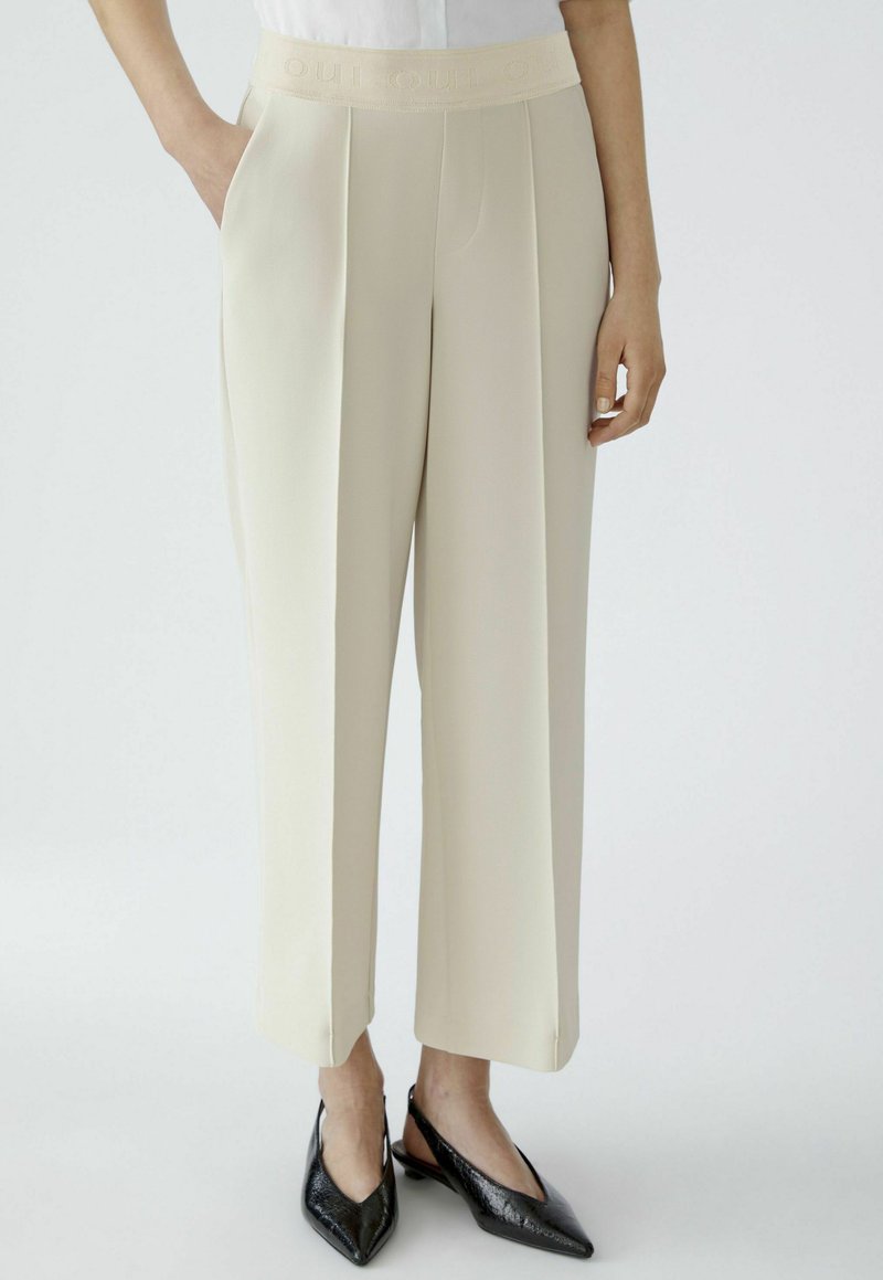 Beige, wide-leg trousers with a textured waistband featuring the word "OU" embossed. Designed with front pleats and side pockets.