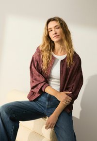 Cazadora bomber burdeos con patrón texturizado, combinada con una blusa blanca de canalé y vaqueros azules de cintura alta. Modelo sentado sobre una superficie de color claro.