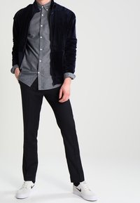 Navy sametová zipová bunda, šedá košile na knoflíky, černé kalhoty, bílé tenisky s černými akcenty. Jednoduchý design, hladká textura a volný střih.