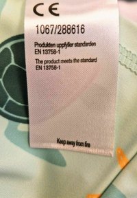 Etikett på tyg med skötselråd, CE-märkning och standarddetaljer. Ljus färg med mjuk textur, tryckt sköldpadda och accentdetaljer.