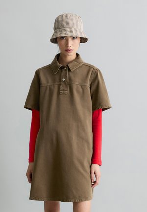 Jeune femme portant une robe marron à manches courtes sur un chemisier rouge à manches longues et un chapeau seau beige à carreaux, debout devant un fond uni.