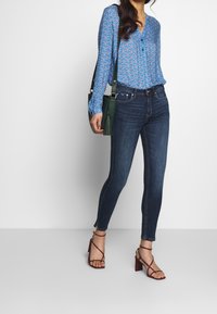 Blå mönstrad blus med långa ärmar, i kombination med mörkblå snäva jeans och bruna sandaler med klack och remmar. Detalj av en grön axelväska synlig.