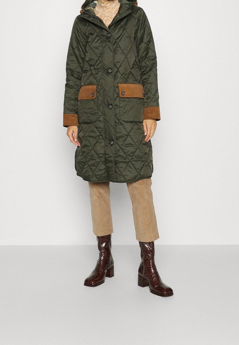 Personne portant un long manteau matelassé vert foncé avec des accents en daim marron, un pantalon beige et des bottes à talons brillantes marron foncé.