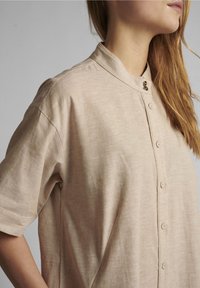 Beżowa koszula na krótki rękaw z guzikami i stójką, z delikatną teksturą i dekoracyjnym detalem guzika na kołnierzyku.