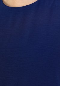 Tessuto blu con una consistenza liscia, caratterizzato da un colletto rotondo e una leggera lucentezza. Nessun motivo visibile o dettagli in metallo.