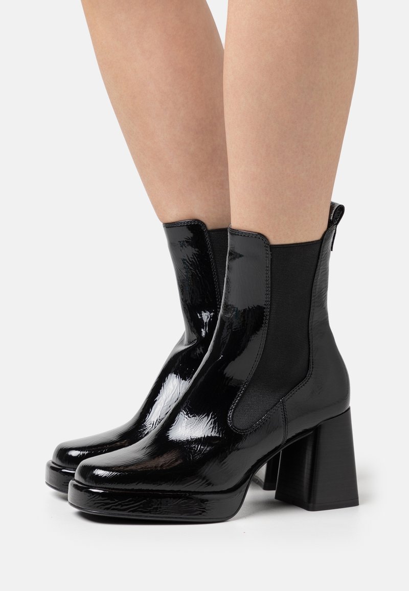 Bottes chevilles en cuir verni noir brillant avec talons compensés portées sur des jambes nues sur un fond clair uni.
