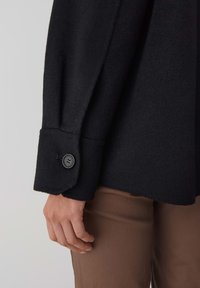 Chemise noire en laine à boutons avec une texture, présentant un poignet arrondi et un unique bouton noir, associée à un pantalon ajusté marron.