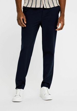 Man die marineblauwe rechte broek en witte sneakers draagt, staand tegen een effen achtergrond.