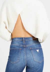Weißer, gerippter Pullover mit tiefem Rückenausschnitt, kombiniert mit blauen Denim-Jeans, die auf der Gesäßtasche ein Dreieckslogo aufweisen.