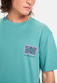 Tyrkysové bavlněné tričko s krátkým rukávem, s růžovým a černým grafickým logem na levé části hrudníku. Kulatý výstřih s jemnou texturou.