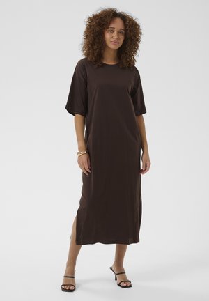 Femme aux cheveux bouclés portant une robe midi ample marron foncé à manches courtes et des sandales à talons noires, debout sur fond blanc.