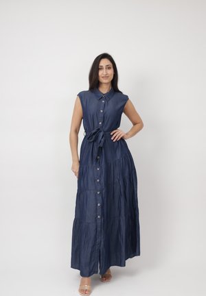 Vrouw staand met hand op heup, gekleed in een mouwloze, donkerblauwe maxi-jurk met knopen en een tailleceintuur, en doorzichtige hakken op een effen witte achtergrond.