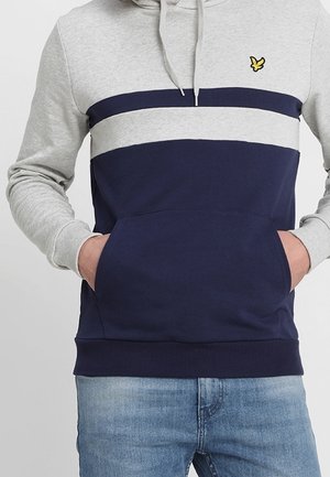 Sudadera con capucha tipo pulóver en gris y azul marino, con rayas horizontales, un bolsillo frontal y un pequeño logo amarillo en el pecho. Textura de tela suave.