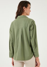 Green long-sleeve shirt made of lightweight fabric, featuring a collared neckline, buttoned cuffs, and a curved hem. Back pleat detail visible.