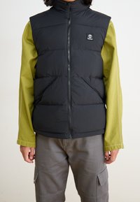 Czarna ocieplana kamizelka z zamkiem błyskawicznym i wysokim kołnierzem, noszona na zielonej koszuli z długim rękawem. Posiada dwie przednie kieszenie oraz naszywkę z logo.