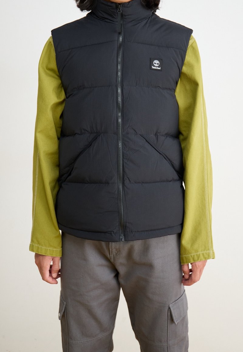 Czarna ocieplana kamizelka z zamkiem błyskawicznym i wysokim kołnierzem, noszona na zielonej koszuli z długim rękawem. Posiada dwie przednie kieszenie oraz naszywkę z logo.