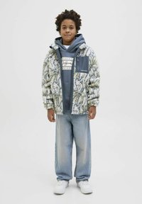 Giacca esterna fuzzy e patterned di un cremisi chiaro e verde, indossata sopra un hoodie blu con logo; abbinata a jeans azzurri e sneakers bianchi.