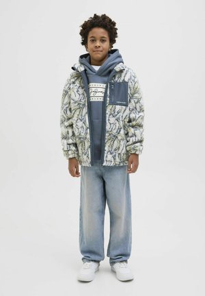 Giacca esterna fuzzy e patterned di un cremisi chiaro e verde, indossata sopra un hoodie blu con logo; abbinata a jeans azzurri e sneakers bianchi.