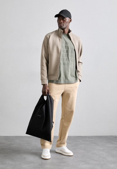 Giacca bomber beige testurizzata, t-shirt grafica verde oliva, pantaloni della tuta beige, borsa nera e sneakers bianche. Design semplice e moderno.