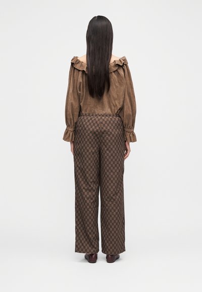 Kvinne med langt mørkt hår står med ryggen til, iført en brun skulderfri topp og mønstrede vide bukser med mørke sko.