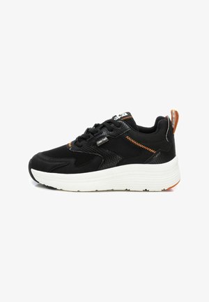 Zapatilla deportiva negra con parte superior de malla y sintético, suela gruesa blanca, costuras de acento naranjas y tab de talón con marca. Paneles texturizados visibles.
