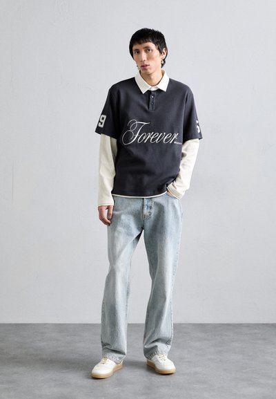Polo de manga corta gris oscuro con el texto "Forever" y el número 9, superpuesto a una camiseta de manga larga blanca; combinado con jeans de color azul claro y zapatillas.
