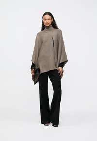 Καφέ πλεχτό πόντσο με ψηλό γιακά, ανοιχτό μπροστά και ασύμμετρο τελείωμα. Συνδυάζεται με μαύρες παντελόνες σε φαρδύ στιλ και μια μικρή μαύρη τσάντα.