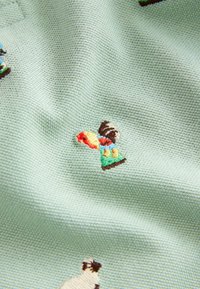 Tissu brodé avec une base vert menthe, présentant des motifs colorés de personnes et d'animaux, mettant en valeur une couture fine et une texture douce.