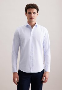 Camicia bianca a maniche lunghe con righe verticali, colletto button-down e orlo curvo. Tessuto liscio con un leggero lucido, indossata con pantaloni scuri.