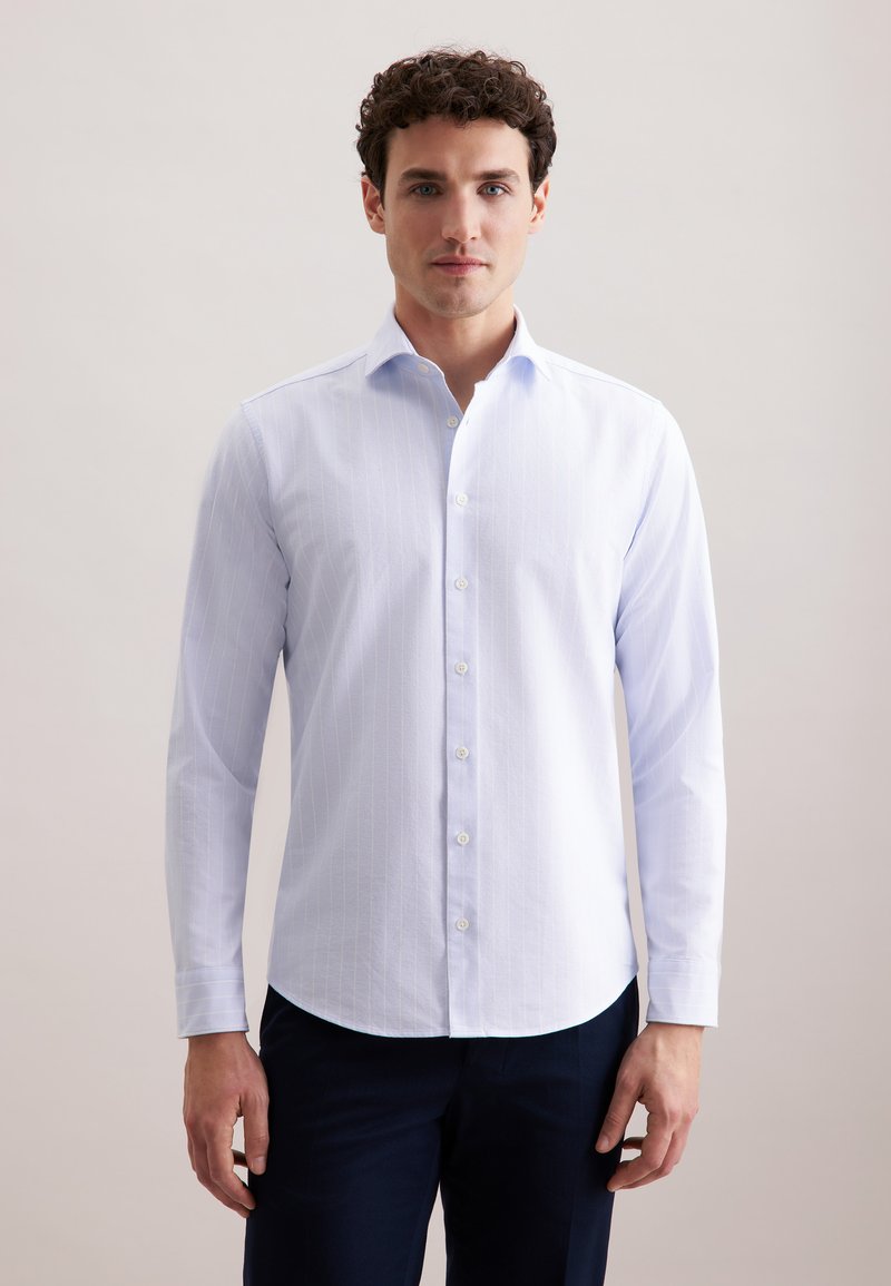 Camicia bianca a maniche lunghe con righe verticali, colletto button-down e orlo curvo. Tessuto liscio con un leggero lucido, indossata con pantaloni scuri.