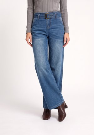 Femme portant un jean bleu taille haute à jambes larges avec une ceinture assortie, associé à un pull gris côtelé et des bottes marron à talons.