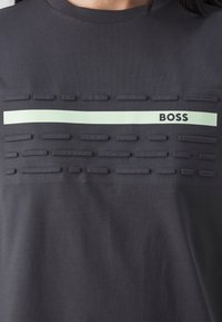Tmavě šedá košile s vyvýšenými obdélníkovými textilními detaily a horizontálním světle zeleným pruhem s nápisem "BOSS" na hrudi.