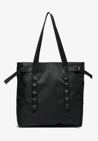 Black tote bag made from durable fabric, featuring two long handles, adjustable straps, and a front pocket. Textured surface with minimal branding.