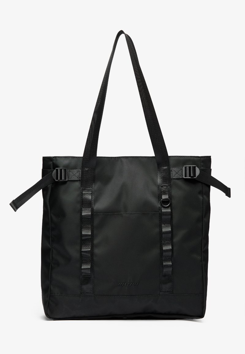 Black tote bag made from durable fabric, featuring two long handles, adjustable straps, and a front pocket. Textured surface with minimal branding.