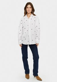 Camicia bianca con motivi a cuore rossi, maniche lunghe e colletto standard; abbinata a jeans blu scuro e stivali a punta marroni.